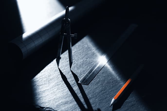 Drawing office instruments on a dark slate bench, crossed by a narrow shaft of hard light: spring dividers throwing a long shadow, a steel rule catching a bright specular streak, a rolled drawing receding into shadow and an orange pencil lying at the edge of the light.